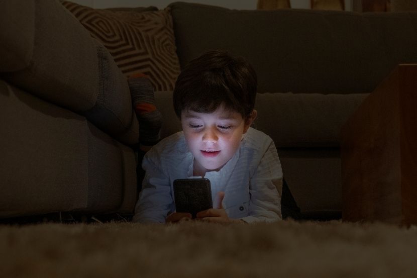 superexposição Criança deitada no chão, olhando atentamente para um celular com a tela iluminada, em ambiente interno, representando o uso excessivo de dispositivos eletrônicos.
