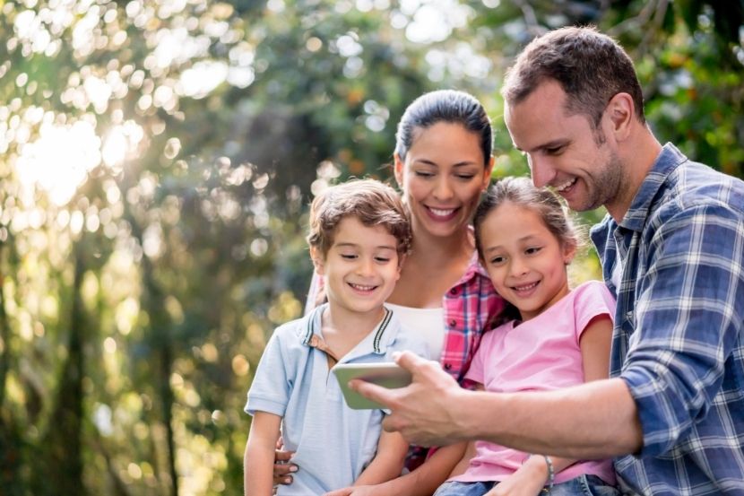 superexposição Família sorridente, incluindo um pai, mãe e dois filhos, visualizando conteúdo em um celular ao ar livre, em meio à natureza, demonstrando união e interação digital.