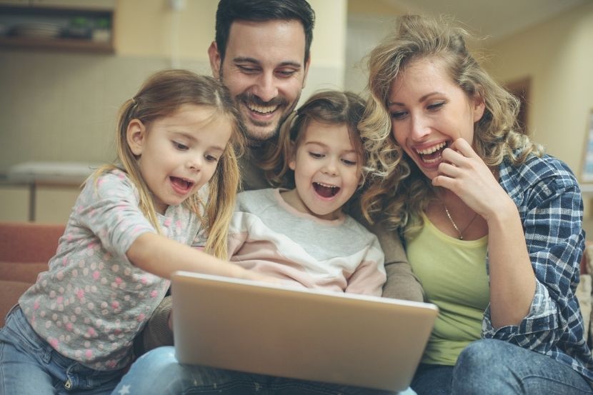 superexposição Família composta por pai, mãe e duas filhas, sorrindo enquanto assistem algo em um laptop, em um ambiente doméstico aconchegante.