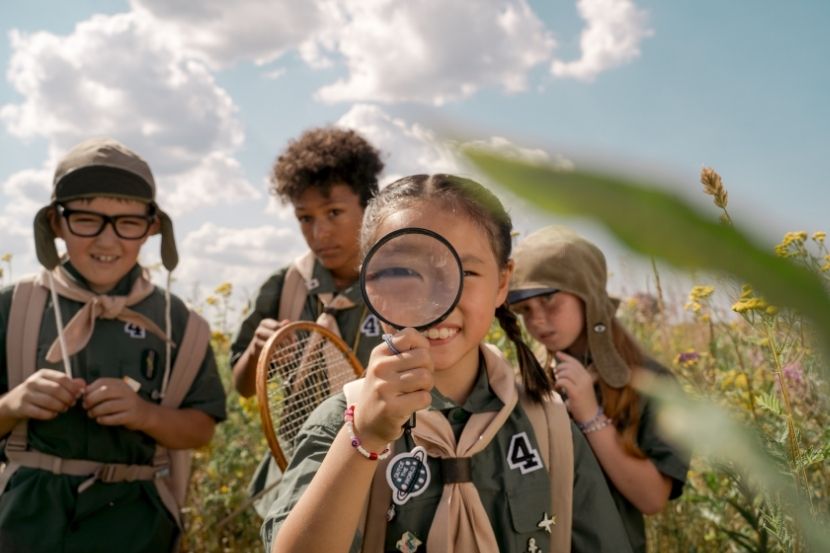 rianças escoteiras, sorrindo e segurando equipamentos de exploração, como uma lupa e rede, enquanto participam de uma atividade ao ar livre, explorando a natureza.