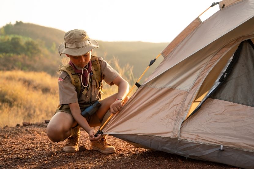 Um jovem escoteiro ajustando a entrada de uma barraca em um acampamento ao ar livre, com o cenário de montanhas e campos ao fundo, durante o entardecer.