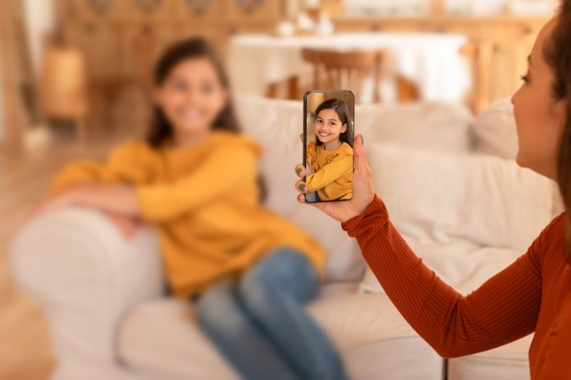 Mulher tirando uma foto da filha, com um smartphone, enquanto ela sorri na frente de um sofá. A criança está usando uma blusa amarela, enquanto o fundo está desfocado.