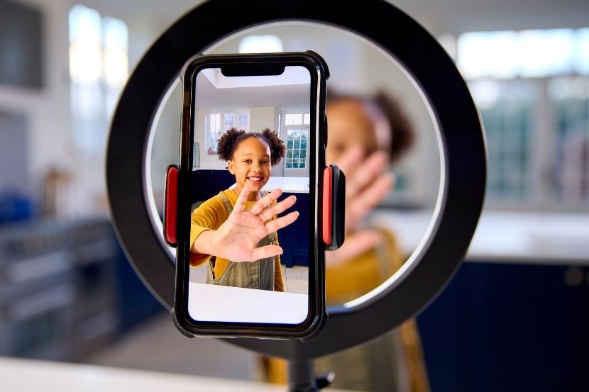 Criança sorrindo e fazendo um gesto de aceno para a câmera, com um smartphone focado nela, enquanto a câmera está posicionada em um aro de luz. O ambiente ao fundo é uma cozinha com luz natural.