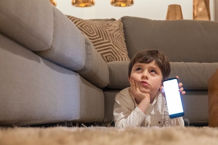 Menino deitado no chão, olhando para a tela de um smartphone em sua mão, com expressão pensativa, em um ambiente de sala de estar com móveis e iluminação suave.