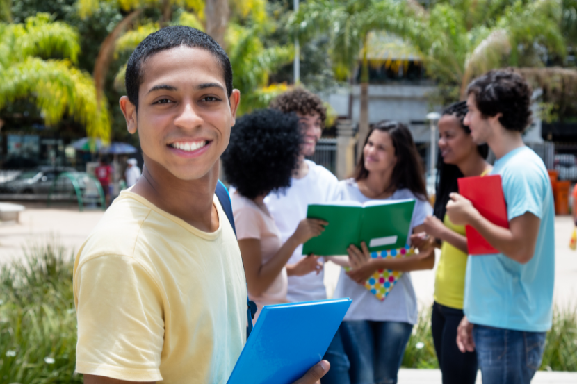 estudante sorrindo com caderno na mão e outros alunos no fundo