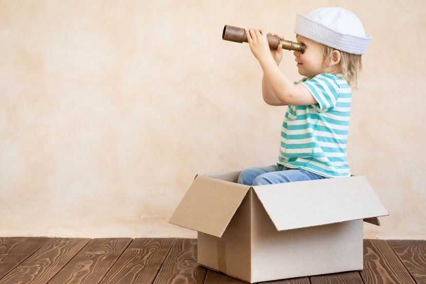 Criança usando um chapéu de marinheiro e olhando através de um telescópio improvisado feito de papelão. Ela está sentada em uma caixa, criando um cenário de brincadeira imaginativa.