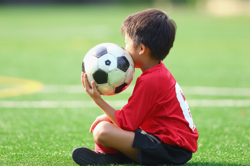 futebol e aprendizado infantil criança sentada com bola na mão e uniforme de futebol
