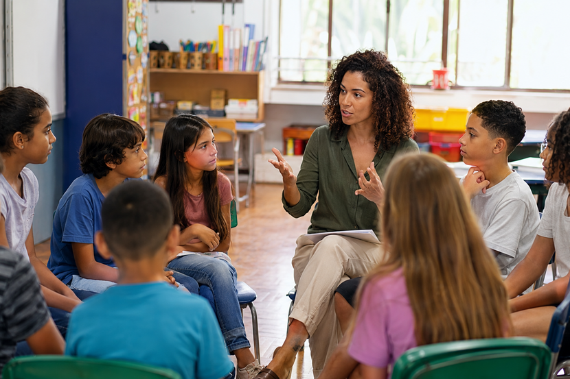 escola laica Professora e estudantes conversam em sala de aula diversa, representando o ambiente de uma escola laica.